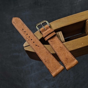 Puede incluir: Una correa de reloj de cuero marrón claro con una superficie texturizada y costuras en contraste. La correa tiene una hebilla plateada y se muestra sobre una superficie de madera. Un accesorio clásico.