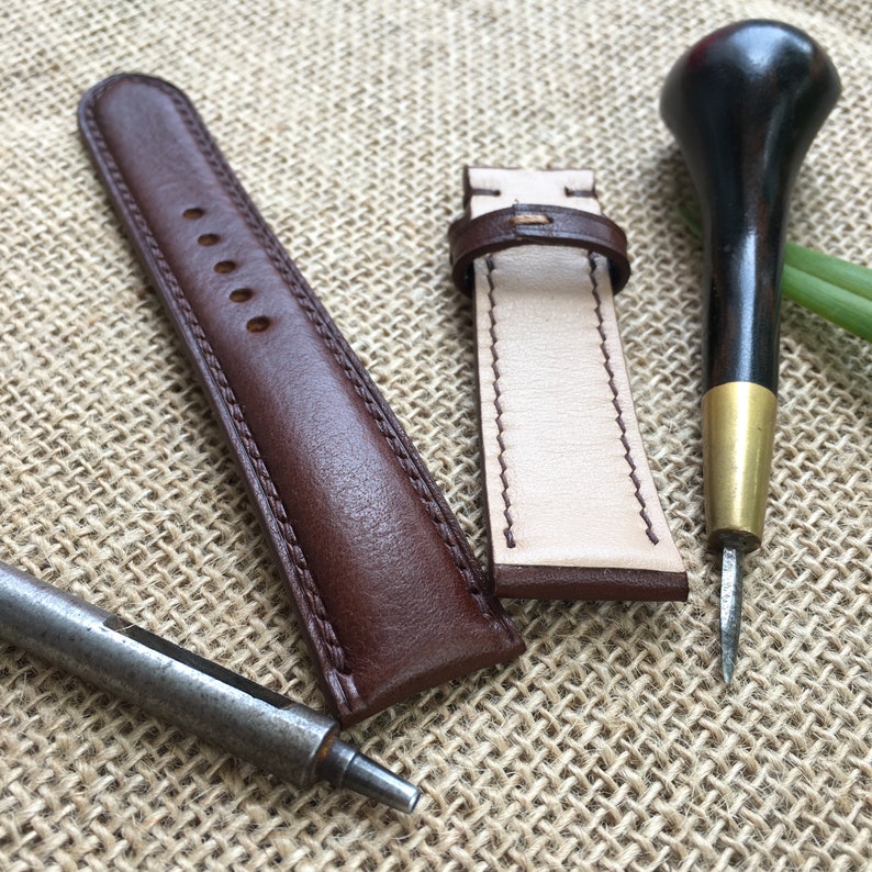 May include: Two brown leather watch straps, one with white stitching and one with brown stitching, are laid out on a brown burlap fabric. A small awl and a wood and brass tool are also on the fabric.
