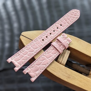 Puede incluir: Correa de reloj rosa claro, con textura de piel de cocodrilo y costuras blancas. La correa es de dos piezas, con agujeros de ajuste visibles. La correa se muestra sobre una superficie de madera.