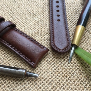 May include: Two brown leather watch straps with stitching detail, lying on a burlap surface. A small metal tool is also visible.