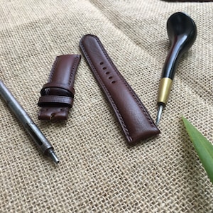 May include: Two brown leather watch straps and a leather tool for crafting watch straps on a brown burlap background.
