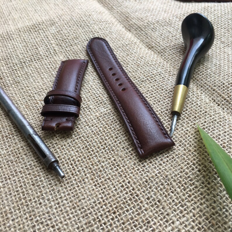May include: Two brown leather watch straps and a leather tool for crafting watch straps on a brown burlap background.