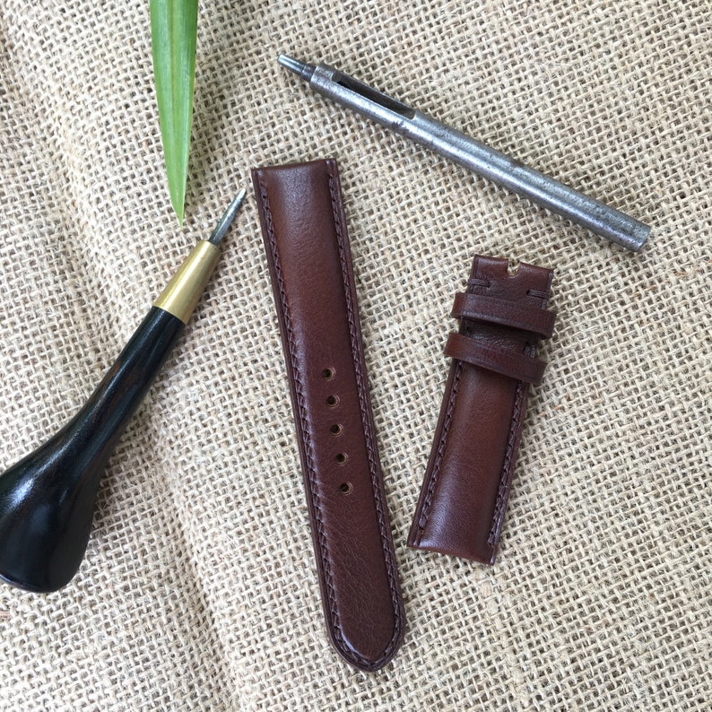 May include: Two brown leather watch straps with stitching detail, a leather awl, and a metal hole punch on a burlap background.