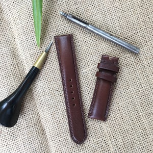 May include: Two brown leather watch straps with stitching detail, a leather awl, and a metal hole punch on a burlap background.