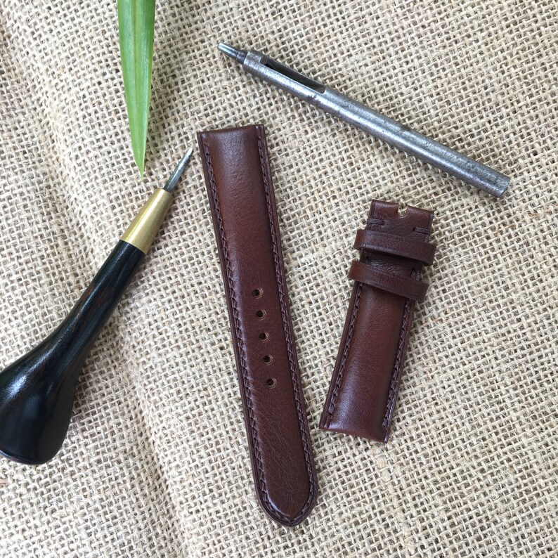 May include: Two brown leather watch straps with stitching detail, a leather awl, and a metal hole punch on a burlap background.