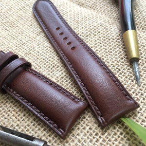 May include: Two brown leather watch straps with stitching detail. The straps are laid out on a brown burlap fabric background.