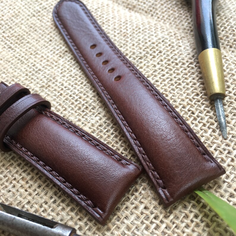 May include: Two brown leather watch straps with stitching detail. The straps are laid out on a brown burlap fabric background.