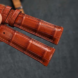 May include: Close-up of two brown leather watch straps with a crocodile skin texture. The straps are a warm, reddish-brown color and feature detailed stitching along the edges. The watch straps are designed to attach to a watch face.
