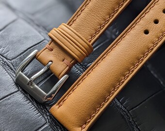 May include: A close-up of a tan leather watch strap with a silver buckle. The strap has a stitched edge and a textured surface.