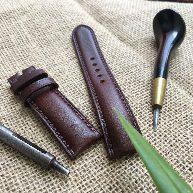 May include: Two brown leather watch straps with stitching, one with a buckle, and a small black tool on a burlap background.