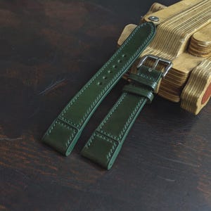 Op de afbeelding: Een donker bosgroene lederen horlogeband met ton-sur-ton stiksels, een gladde afwerking en een zilverkleurige metalen gesp. De band bestaat uit twee delen met meerdere verstelgaatjes en twee lederen lussen om de horlogeband vast te zetten.