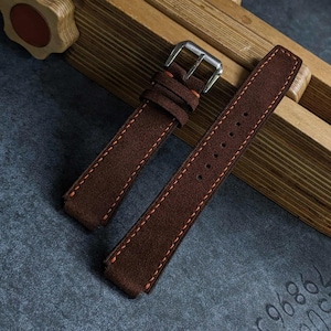 May include: A dark brown suede watch strap featuring contrasting orange stitching along the edges. The strap includes a silver-toned metal buckle and a matching sliding keeper loop, with a series of punched holes for adjustable sizing.
