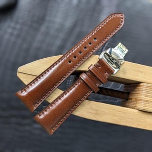 Puede incluir: Una correa de reloj de cuero marrón con costuras a tono, una serie de orificios de ajuste circulares y un cierre desplegable de metal plateado pulido. La correa consta de dos piezas, con el extremo de la hebilla mostrando dos trabillas.