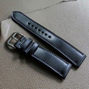 Könnte beinhalten: Zwei schwarze Lederarmbänder mit Nähten. Das obere Armband hat eine silberne Schnalle und ist in der Mitte gefaltet. Das untere Armband ist länger und hat eine silberne Schnalle.