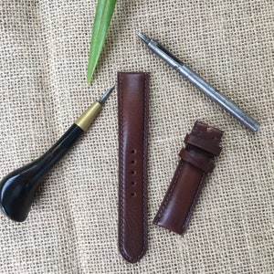 May include: Two brown leather watch straps with stitching and a leather punch tool. The straps are laid out on a burlap background.