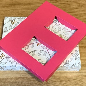 May include: A pink cardboard box with two rectangular cutouts. The box is placed on top of a smaller box with a white background and a pattern of brown leaves.
