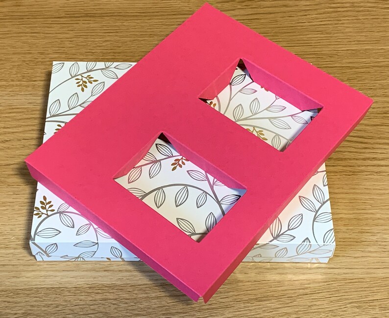 May include: A pink cardboard box with two rectangular cutouts. The box is placed on top of a smaller box with a white background and a pattern of brown leaves.