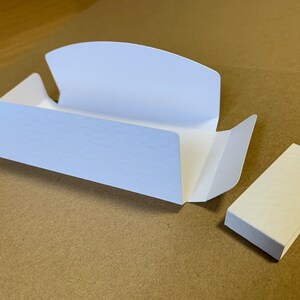 May include: A white cardboard box with a flap that folds over to create a small, rectangular box. The box is open and the flap is folded back. There is a separate white cardboard piece next to the box.