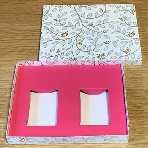 May include: A white box with a floral pattern and a pink interior. The box has two rectangular cutouts in the pink interior.