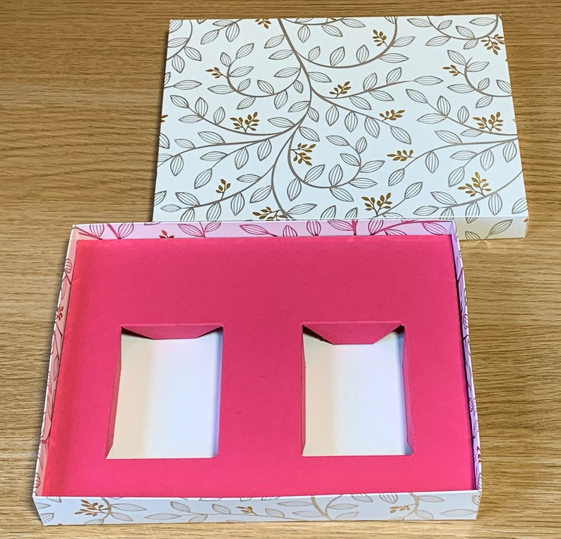 May include: A white box with a floral pattern and a pink interior. The box has two rectangular cutouts in the pink interior.