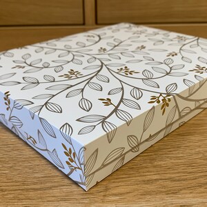 May include: A white gift box with a gold floral pattern. The box is closed and sitting on a wooden surface.