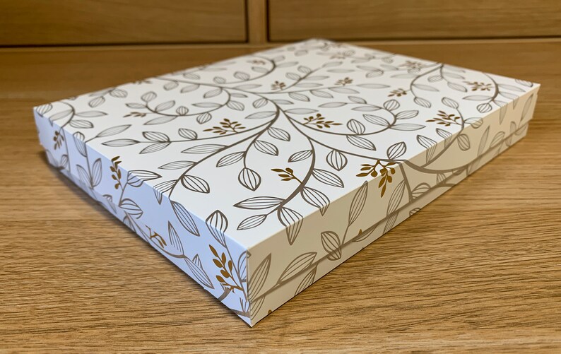 May include: A white gift box with a gold floral pattern. The box is closed and sitting on a wooden surface.
