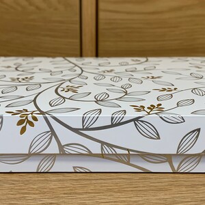 May include: White gift box with a gold and gray floral pattern. The box is closed and the lid is facing the camera.