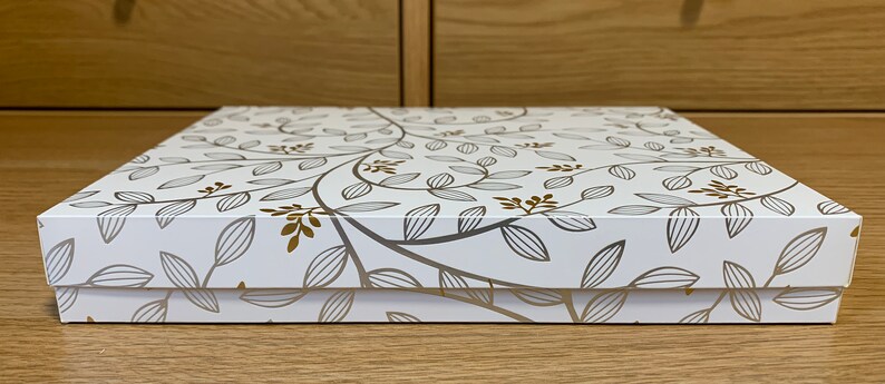 May include: White gift box with a gold and gray floral pattern. The box is closed and the lid is facing the camera.