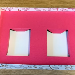 May include: A white box with a floral pattern on the outside and a pink interior. The box has two rectangular cutouts in the pink interior.