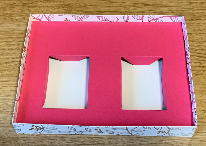 May include: A white box with a floral pattern on the outside and a pink interior. The box has two rectangular cutouts in the pink interior.