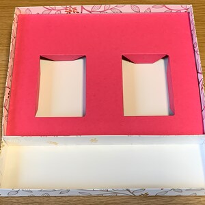 May include: A pink and white gift box with a floral pattern. The box has two rectangular cutouts in the top section and a white interior.