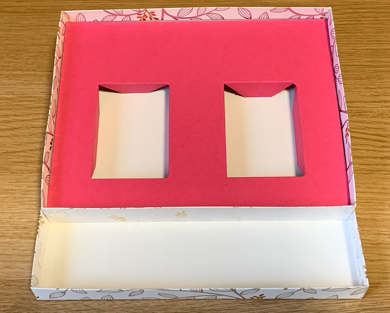 May include: A pink and white gift box with a floral pattern. The box has two rectangular cutouts in the top section and a white interior.