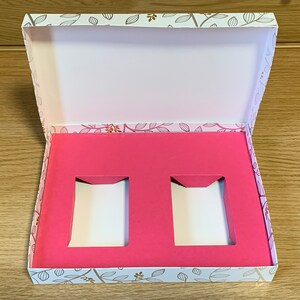 May include: An open white box with a floral pattern on the outside. The inside of the box is lined with pink foam and has two rectangular cutouts.
