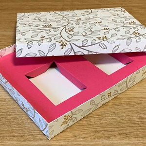 May include: A white gift box with a floral pattern on the lid and a pink interior with two rectangular cutouts. The box is open and the lid is propped up.