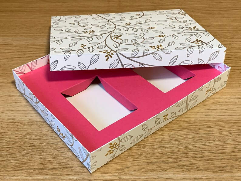 May include: A white gift box with a floral pattern on the lid and a pink interior with two rectangular cutouts. The box is open and the lid is propped up.