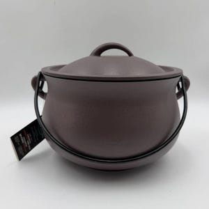 May include: A dark brown cast iron pot with a matching lid and a black metal bail handle. The pot features a rounded body and two side handles. A rectangular black product tag with white text is attached to one of the side handles.