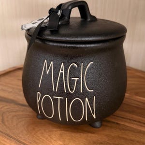 May include: A black ceramic cauldron-shaped jar with a lid and three small feet. The front features the white text MAGIC POTION in a handwritten font. A black ribbon is tied to the handle, holding a small white tag with a skull illustration.