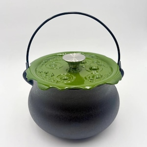 May include: A round, dark gray cast iron pot with a matte finish and a black metal handle. The pot features a glossy, olive green lid with a scalloped edge and a raised, circular bubble pattern. A silver-toned, conical knob sits at the center.