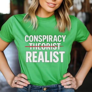 以下が含まれることがあります： 「CONSPIRACY THEORIST」という文字が打ち消され、「REALIST」という文字がその下に書かれた緑色のTシャツです。