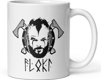 Puede incluir: Taza de cerámica blanca con una ilustración en negro y gris de un guerrero vikingo, hachas cruzadas y la palabra "FLOKA" en letras estilizadas. El diseño incluye una cara detallada con barba y pintura de guerra.
