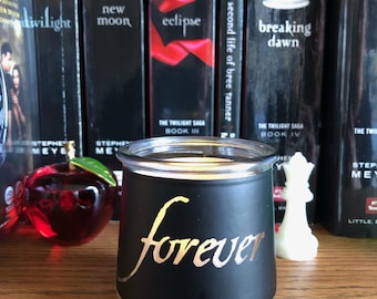 May include: A black glass candle holder with the word "forever" in gold script. The candle holder is in front of a row of books, including "Twilight" and "Breaking Dawn". A decorative red apple is to the left.