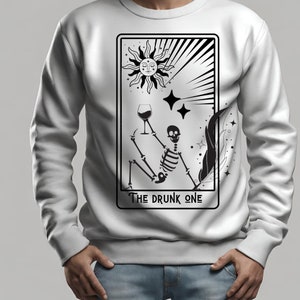 May include: White sweatshirt with a black and white illustration of a skeleton holding a glass of wine. The illustration is in the style of a tarot card and says "The Drunk One" at the bottom.