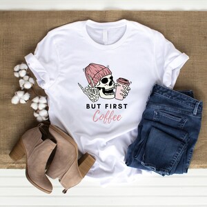 May include: White t-shirt with a pink and black skeleton wearing a pink beanie and holding a coffee cup. The text "But First Coffee" is printed below the skeleton.