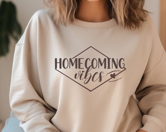 Puede incluir: Una sudadera beige con el texto "HOMECOMING vibes" en forma de diamante con un pequeño corazón dentro de un gráfico de fútbol.