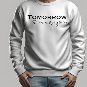 以下が含まれることがあります： 白いスウェットシャツに、黒い文字で「TOMORROW ♥ needs you」と書かれています。