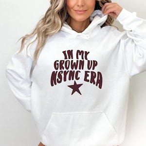 Op de afbeelding: Witte hoodie met een kastanjebruine afbeelding met de tekst "In My Grown Up NSYNC Era" met een kastanjebruine ster.