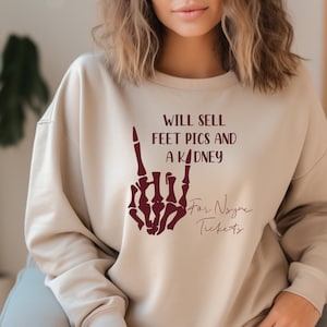 May include: A beige sweatshirt with a maroon graphic of a skeleton hand making a rock and roll sign. The text "Will sell feet pics and a kidney for Nsync tickets" is printed on the sweatshirt.