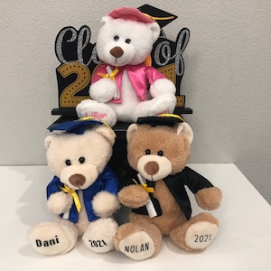 Pu&ograve; includere: Tre orsacchiotti che indossano cappelli e toge da laurea. Gli orsi sono seduti su un cartello di legno nero che dice "Classe del 2021". Gli orsi si chiamano Dani, Nolan e Lillian.