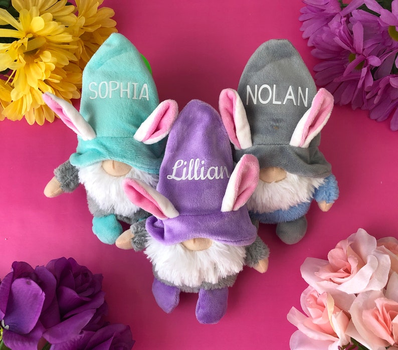 Puede incluir: Tres gnomos de peluche con orejas de conejo y nombres personalizados: Sophia, Nolan y Lillian. Los gnomos llevan sombreros de colores pastel y tienen barbas blancas. Los gnomos est&aacute;n sentados sobre una superficie rosa.