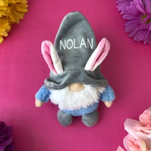 Puede incluir: Un gnomo de peluche gris con una camisa azul y barba blanca que lleva un gorro de conejo gris con orejas rosas. El gorro tiene el nombre "NOLAN" bordado.